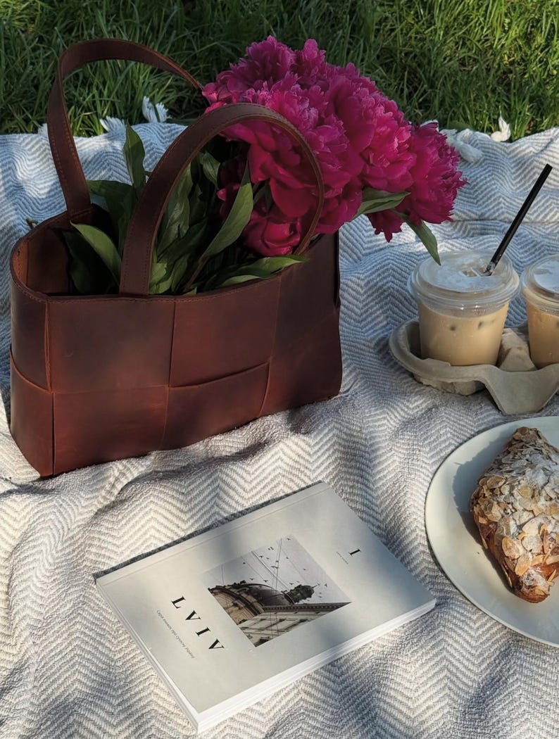 Puede incluir: Escena de picnic con un bolso tote de cuero marr&oacute;n lleno de peon&iacute;as rosas. Dos caf&eacute;s helados con pajitas, un cruas&aacute;n y un libro con la palabra "LVIV" sobre una manta blanca.