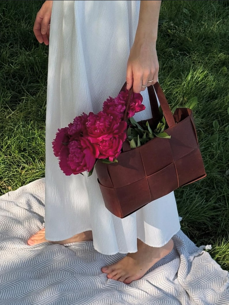 Puede incluir: Un bolso tote de cuero marr&oacute;n tejido, lleno de vibrantes peon&iacute;as rosas. El bolso tiene forma rectangular con un asa. La persona lleva un vestido blanco y est&aacute; de pie sobre una manta en la hierba.