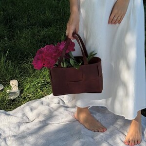 Puede incluir: Un bolso tote burdeos tejido lleno de vibrantes peon&iacute;as rosas y hojas verdes. El bolso tiene dos asas y lo sostiene una persona que lleva un vestido blanco. La escena est&aacute; ambientada al aire libre sobre una manta y hierba.