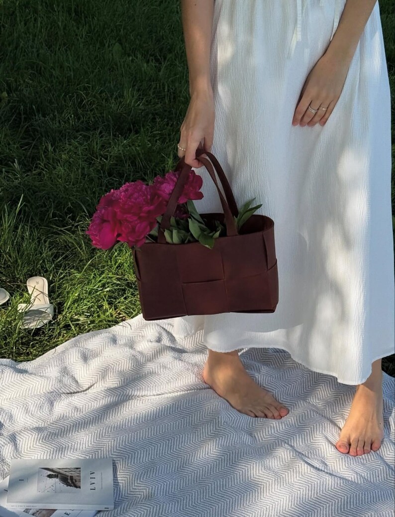Puede incluir: Un bolso tote burdeos tejido lleno de vibrantes peon&iacute;as rosas y hojas verdes. El bolso tiene dos asas y lo sostiene una persona que lleva un vestido blanco. La escena est&aacute; ambientada al aire libre sobre una manta y hierba.