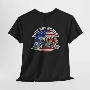 May include: Black t-shirt featuring a graphic of a jet plane with flames, set against a distressed American flag design. The text "Ugly But Honest" is arched above the image. The shirt is made of a soft material.