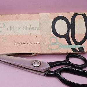 May include: Vintage pinking shears with a silver blade and black handles, resting in front of a worn cardboard box. The box reads "Pinking Shears" and "Cutlers Guild, Limited."