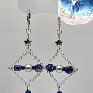 May include: Silver dangle earrings with star-shaped studs, diamond-shaped blue and silver beadwork, and crescent moon charms. The earrings have blue faceted beads and silver chain links. The earrings are hanging from a white tag.