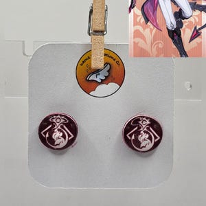 May include: A pair of round, maroon-colored stud earrings with a white and black graphic design. The earrings are displayed on a white card, held by a wooden clip. The card also features a logo with text.