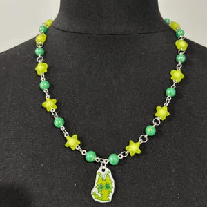 May include: A green and yellow beaded necklace with a pendant. The necklace features green beads, yellow star-shaped beads, and a pendant with a cartoon creature design. The necklace is displayed on a black mannequin.