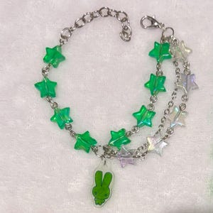 May include: A multi-strand silver-tone metal chain bracelet featuring translucent green star-shaped beads and iridescent clear star beads. A small, flat, green rabbit-shaped charm hangs from the center of the bracelet, which includes a lobster claw clasp.