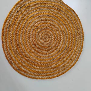 Puede incluir: Una alfombra redonda de yute trenzado con un diseño en espiral. La alfombra presenta bandas alternas de color beige natural y amarillo dorado. Una cubierta de suelo decorativa.