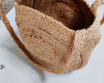 May include: A natural-toned jute tote bag with braided handles. The bag's body is woven in a spiral pattern, creating a textured surface. The handles are thick and sturdy, designed for carrying. The bag is open at the top, revealing the interior.