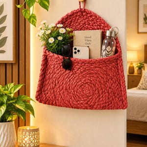 May include: A red woven wall-hanging storage pocket featuring a triangular flap and a loop for hanging. The pocket contains a small bouquet of white daisies, a pair of black sunglasses, a smartphone, and a book with the text Good Vibes Only.