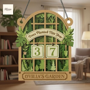Könnte beinhalten: Garten-Holzschild mit einem geschichteten Wald-Design. Das Schild zeigt "Trees Planted This Year" und "37" mit dem Text "Ovilia's Garden" unten. Das Schild hängt an einer Kette.