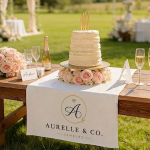 以下が含まれることがあります： Aurelle & Co. Jewelryの黒いロゴが入った白いテーブルランナー。その上には、段重ねの白いケーキ、金色のキャンドル、ピンクのバラの土台が飾られています。テーブルにはシャンパンボトル、グラス2つ、Andersonと書かれた席札が置かれています。