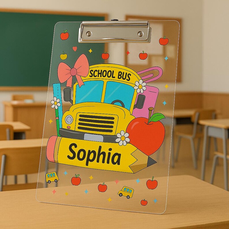 May include: A clear clipboard with a colourful school bus design, including the text "School Bus" and "Sophia". The design incorporates a red apple, pink bow, and other school-related illustrations. The clipboard has a silver metal clip.