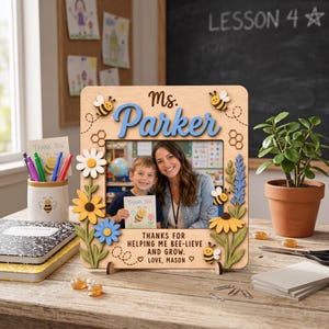 Può includere: Una cornice portafoto in legno chiaro con il nome Ms. Parker intagliato in corsivo blu, decorata con sagome in legno di api, margherite e fiori di campo. La cornice riporta il testo: THANKS FOR HELPING ME BEE-LIEVE AND GROW. LOVE, MASON.