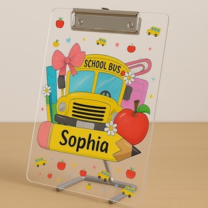 May include: A clear acrylic clipboard with a school bus, apple, and pencil design. The clipboard features the name "Sophia" and is decorated with school-themed illustrations, including apples and school buses.
