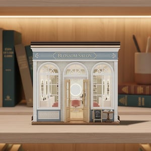 Puede incluir: Maqueta en miniatura de un salón Blossom con fachada azul claro y blanca. El modelo presenta ventanas arqueadas, un interior detallado con sillones de salón y una pequeña pizarra. El nombre del salón se muestra sobre las ventanas.