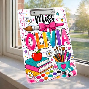 May include: A personalized clipboard featuring the name "Olivia" in colorful letters. The design includes a paintbrush, bow, apple, books, paint palette, and art supplies. The word "Miss" is written above the name. The clipboard has a metal clip at the top.