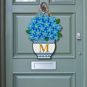 May include: A wooden door hanging featuring a blue hydrangea flower arrangement in a white and black vase with the letter "M". The door decoration is hanging on a teal door with a silver door knocker.