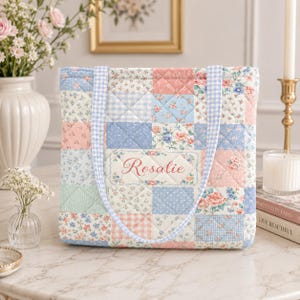 Könnte beinhalten: Eine gesteppte Tragetasche mit einem Patchwork-Design aus Blumen- und Vichy-Karomustern in Pastellrosa, Blau und Weiß. Die Tasche hat zwei hellblau-weiß karierte Henkel und ein zentrales rechteckiges Etikett mit dem Namen Rosalie in roter Schrift.
