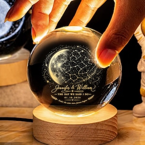 May include: A clear glass sphere with a celestial design, illuminated by a warm light from a wooden base. The sphere features a moon, constellations, and the names "Jennifer & William" with a date. The text reads "THE DAY WE SAID I DO - 01.10.2026".