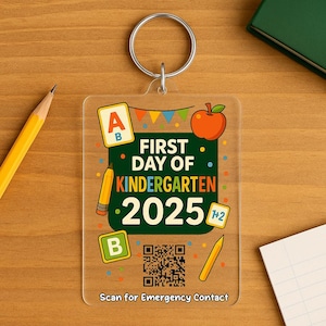 May include: Clear acrylic keyring with a silver ring. The keyring features the text "First Day of Kindergarten 2025" with colourful graphics of an apple, pencils, and the letters A and B. The bottom of the keyring reads "Scan for Emergency Contact."
