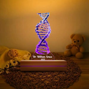 Könnte beinhalten: Ein Nachtlicht in Form einer DNA-Helix mit einem lila-blauen Farbverlauf. Das Licht steht auf einem Holzsockel und trägt den Text "Dr. William Grace 2025". Im Hintergrund ist ein Teddybär zu sehen.