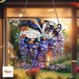 May include: A stained glass cat suncatcher. The cat is sleeping in a flower pot filled with purple wisteria. The cat has orange, black, and white markings. The suncatcher hangs from a silver chain.
