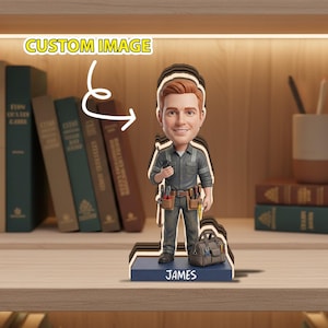 Puede incluir: Figura de un electricista sonriente con uniforme gris, cinturón de herramientas y caja de herramientas. La base es azul con el nombre "JAMES" en blanco. El texto "CUSTOM IMAGE" está en la parte superior.