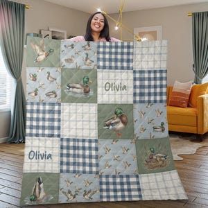 May include: A patchwork quilt featuring duck illustrations and blue gingham patterns. The quilt has the name "Olivia" embroidered on two squares. The color palette includes shades of blue, green, and white, creating a cozy and rustic aesthetic.