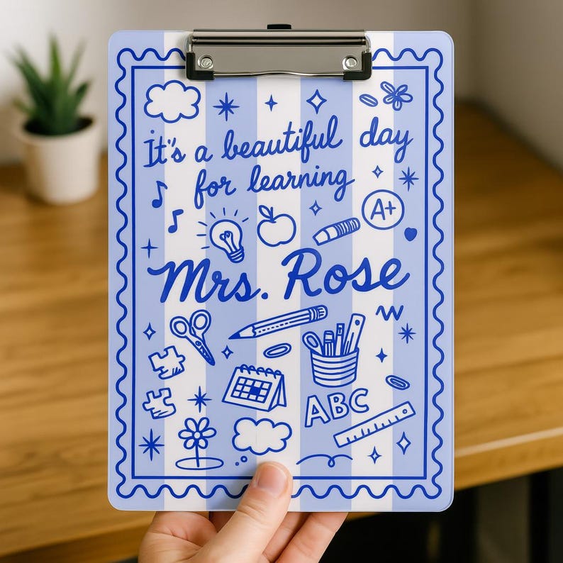K&ouml;nnte beinhalten: Ein blau-wei&szlig; gestreifter Klemmbrett mit Metallclip. Das Klemmbrett zeigt den Text "It's a beautiful day for learning Mrs. Rose" und verschiedene Illustrationen zum Thema Schule in Blau.