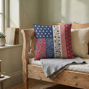 May include: Square decorative pillow with a patchwork design. The pillow features sections with a blue field of white stars, red paisley, blue paisley, floral patterns, red gingham, and red circles with white designs. The pillow is on a wooden bench.
