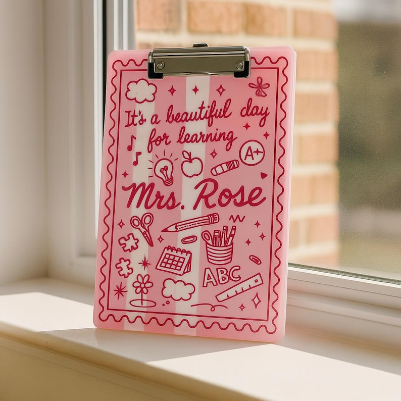 K&ouml;nnte beinhalten: Rosa Klemmbrett mit silbernem Clip, mit dem Text "It's a beautiful day for learning Mrs. Rose" und Illustrationen zum Thema Schule in Rot. Das Klemmbrett hat einen wellenf&ouml;rmigen Rand und vertikale Streifen.