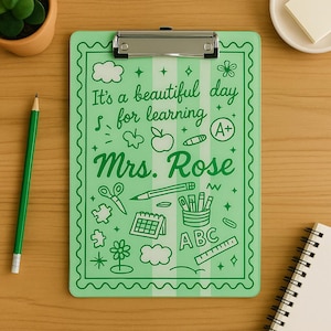K&ouml;nnte beinhalten: Ein gr&uuml;nes Klemmbrett mit silbernem Clip, mit dem Text "It's a beautiful day for learning Mrs. Rose" und Illustrationen zum Thema Schule. Ein gr&uuml;ner Bleistift liegt auf der Holzoberfl&auml;che.