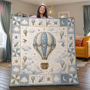 May include: A quilted blanket featuring a large central hot air balloon in shades of blue and cream, surrounded by smaller balloons, clouds, stars, feathers, and birds. The border consists of alternating squares with geometric patterns and motifs.
