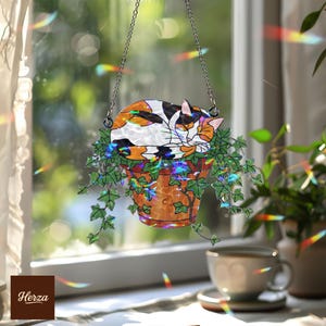 May include: A stained glass suncatcher featuring a sleeping calico cat resting in a terracotta pot with green ivy. The cat has orange, black, and white markings. The suncatcher hangs from a silver chain. The word "Herza" is in the bottom left corner.
