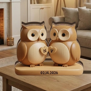 Puede incluir: Dos figuras de búhos de madera con recortes en forma de corazón, cada uno con una letra. Los búhos están sobre una base de madera con la fecha "02.14.2026" grabada. Los búhos tienen ojos grandes y redondos y están en un ambiente doméstico.