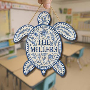 Puede incluir: Cartel de madera con forma de tortuga con un diseño floral azul y blanco. El cartel presenta el texto "THE MILLERS" en una fuente decorativa y está suspendido por una cuerda natural. El cartel es un artículo decorativo.