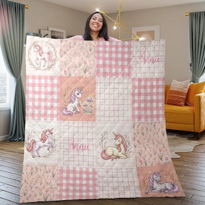 Puede incluir: Una colcha de patchwork rosa y blanca con ilustraciones de unicornios y el nombre "Annie". La colcha tiene una mezcla de patrones florales, cuadros y cuadrículas. Los unicornios se representan en varias poses, rodeados de coronas florales.