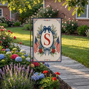May include: A garden flag with a patriotic design. It features the letter "S" in red, surrounded by a floral wreath, two American flags, and blue bows. The flag has a striped background and a navy border.