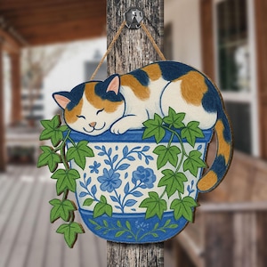 May include: A wooden sign depicting a sleeping calico cat on a blue and white floral planter. The planter has green vines and leaves. The cat has orange, black, and white fur. The sign is suspended from a wooden post with a twine string.