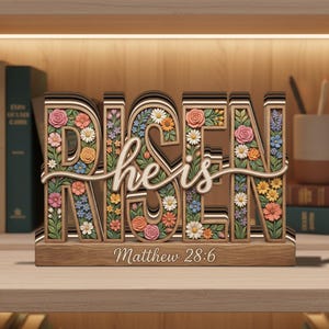 May include: A wooden decorative sign with the word "RISEN" in large, layered letters, filled with colorful flowers. The words "he is" are in a cursive font, and the text "Matthew 28:6" is at the base. The sign is on a wooden shelf.