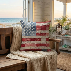 May include: A cushion with a patchwork design resembling the American flag, in red, white, and blue fabrics. It rests on a wooden bench draped with a cream-coloured knitted blanket. The scene is set on a porch overlooking the sea, with a lantern visible.