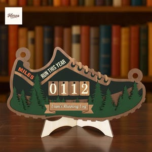Pode incluir: Placa de madeira em forma de tênis de corrida com o texto "RUN THIS YEAR" e "MILES". A placa tem um contador exibindo "0112" e o texto "Liam's Running Log". O tênis é verde e marrom, com um suporte de madeira.