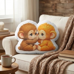 May include: A decorative pillow featuring two cartoon monkeys in shades of brown and orange. The monkeys are sitting close together, with one covering its mouth. The pillow is on a cream-colored couch with a brown knitted throw.