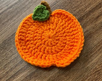 Puede incluir: Un posavasos de ganchillo con forma de calabaza naranja hecho a mano, con una hoja verde y un tallo marrón. El posavasos es redondo y tiene una superficie texturizada, ideal para la decoración otoñal. La calabaza mide unos 10 cm de diámetro.