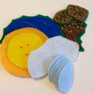 May include: A felt craft set with a yellow sun, blue water, green grass, brown tree, and blue clouds. The sun has a smiling face with red dots for eyes.