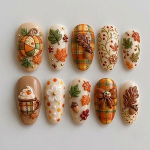 May include: A set of ten oval-shaped press-on nails featuring 3D autumn-themed designs. Decorations include raised pumpkins, acorns, maple leaves, plaid patterns, a mug with whipped cream, polka dots, a brown bow, and small gold metallic beads.