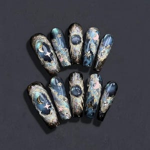 Puede incluir: Un conjunto de diez uñas postizas largas en forma de ataúd. Cada uña presenta un diseño de galaxia azul oscuro y plateado con una luna creciente, estrellas y una gema azul redonda grande. Algunas uñas tienen acentos rosados. Las uñas están dispuestas sobre una superficie gris oscuro.