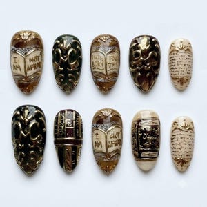 May include: A set of ten almond-shaped press-on nails featuring intricate gold relief designs, including open books with the text I AM NOT AFRAID, ornate scrollwork, and script patterns in shades of black, cream, brown, and deep burgundy.
