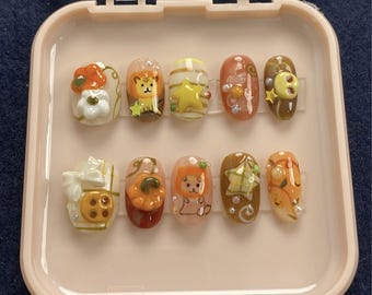 May include: A set of ten press-on nails in a pink case. The nails feature various designs, including a lion, a star, a pumpkin, and floral accents. Colors include white, orange, brown, and yellow. Some nails have pearl embellishments.