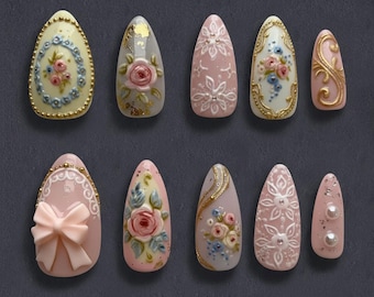 May include: A collection of ten artificial nails in various designs. The nails feature intricate floral patterns, bows, and gold accents on a pink, white, and cream base. Some nails have a matte finish, while others have a glossy appearance.